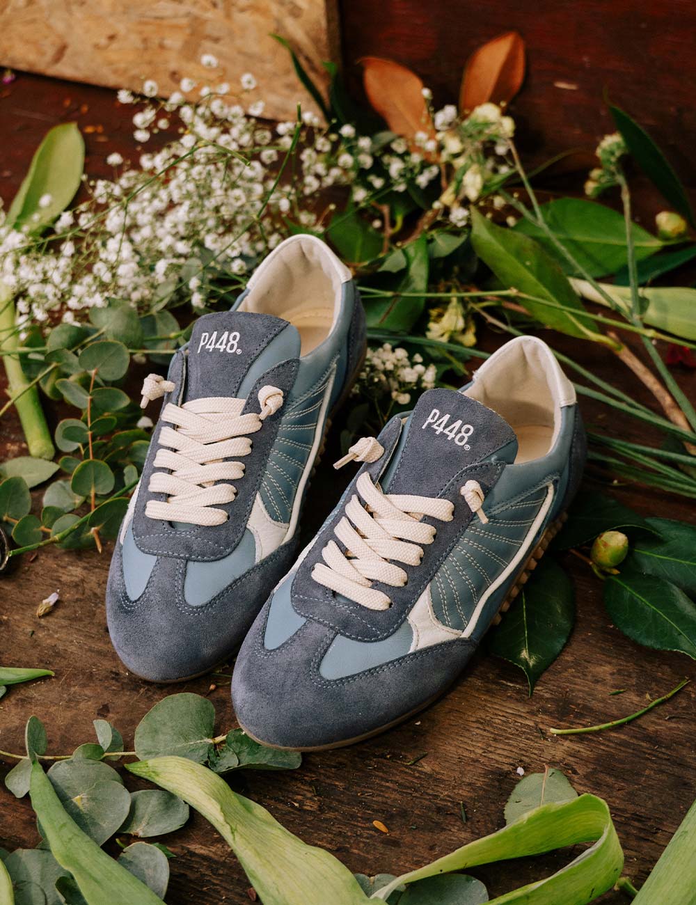 Top-down view of P448 blue leather and blue suede sneakers on a rustic wooden surface, surrounded by eucalyptus leaves and white flowers. Features cream laces, contrast stitching, and a white heel tab.
