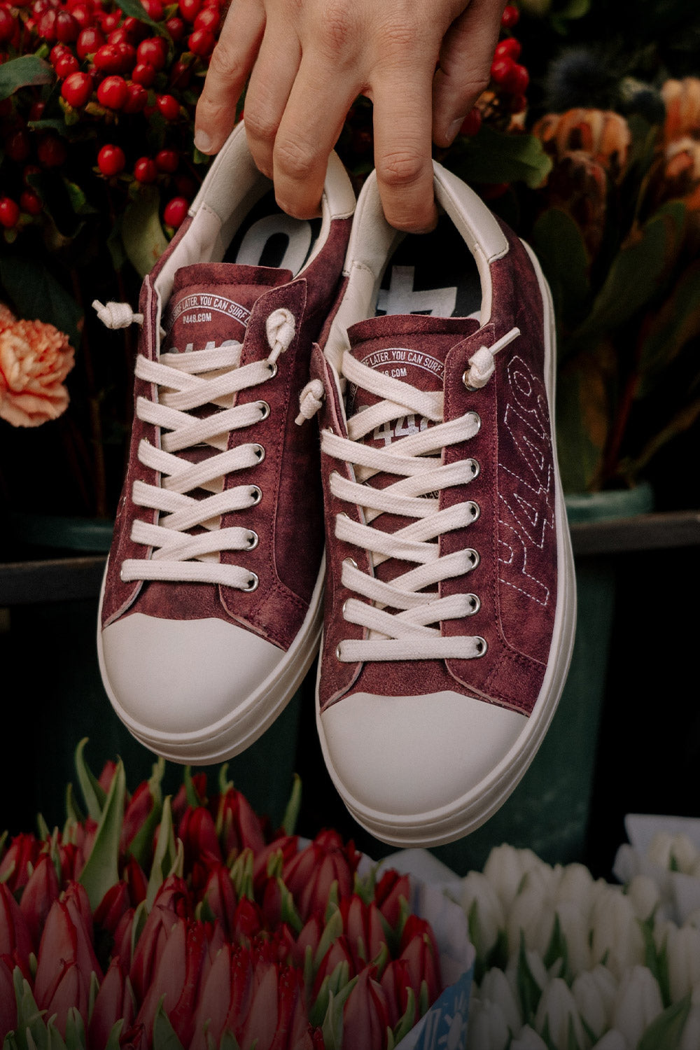 P448 low-top sneaker in burgundy suede with white laces, held by hand in front of an arrangement of fresh flowers and red berries.