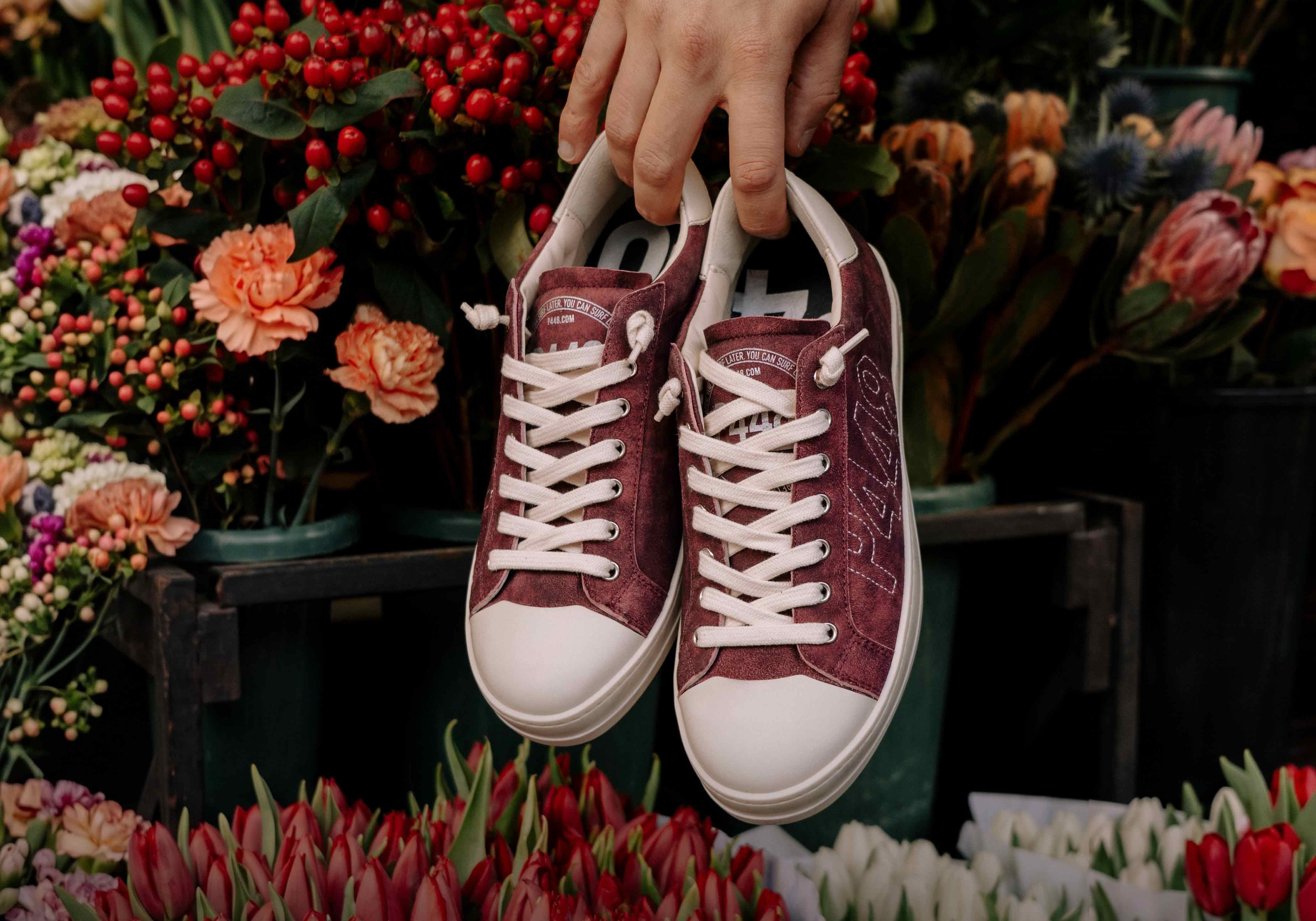 P448 low-top sneaker in burgundy suede with white laces, held by hand in front of an arrangement of fresh flowers and red berries.