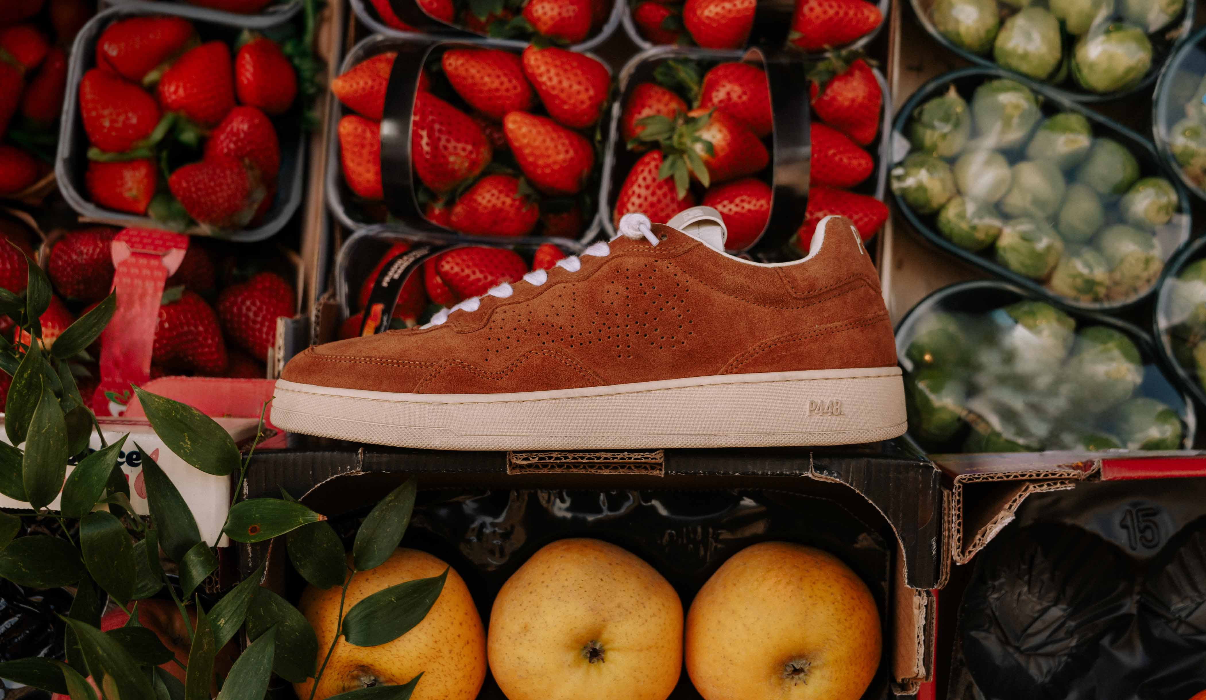 Red suede P448 low-top sneaker featuring white laces and a cream rubber sole with debossed branding, showcased in a vibrant market setting.