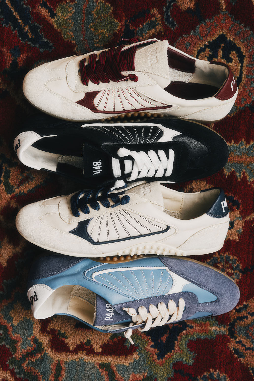 Four pairs of P448 Lisbon designer sneakers in burgundy, black, navy, and blue colorways on a vintage patterned rug.