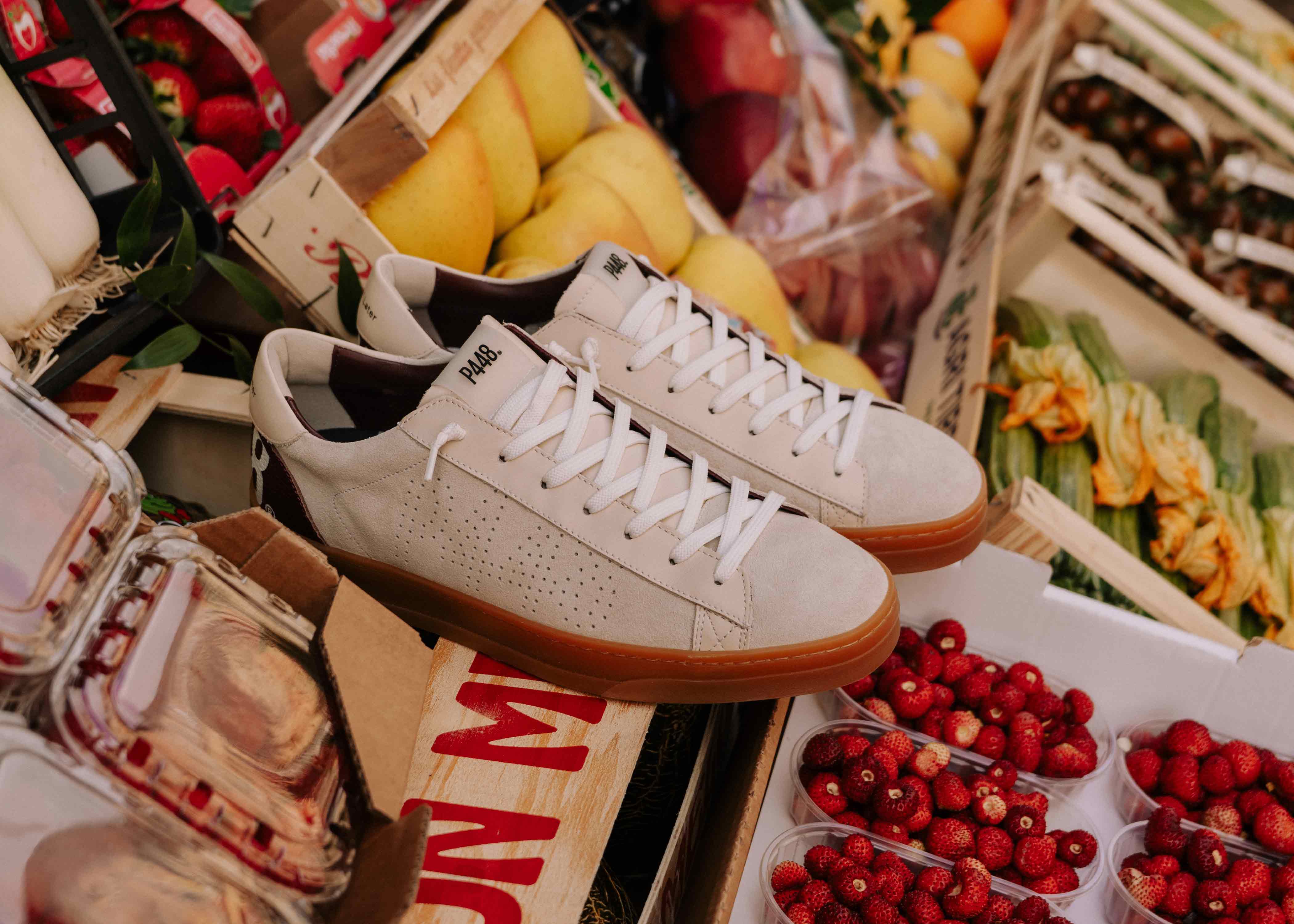 Pair of P448 sneakers in off-white suede with a gum rubber sole, displayed amidst fresh fruit and wooden crates at a market. Features a perforated side logo, white laces, and a dark burgundy heel tab.
